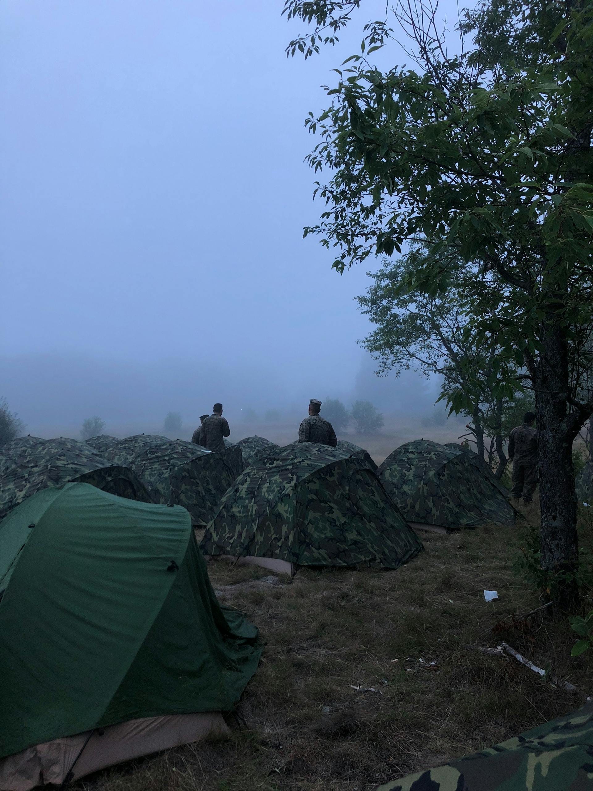 Soldiers stand among tents in a misty field, trees visible nearby